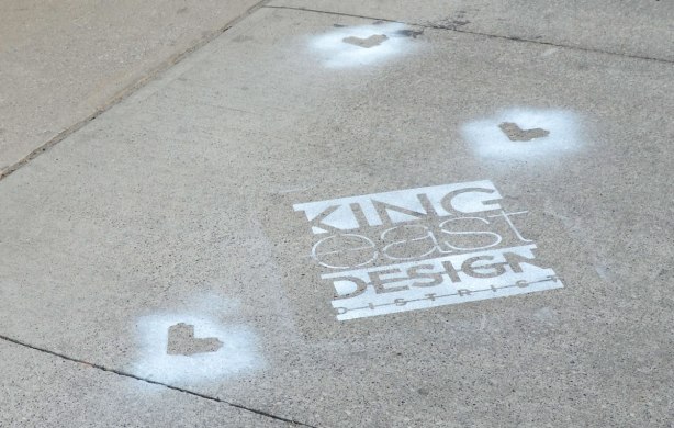 A section of sidewalk. On it is painted the logo for King East Design District. There are also three lovebot stencil shapes spray painted in white on the sidewalk. 