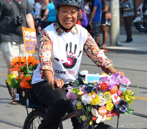 An Asian woman rides her bike in a labour day parade. She is wearing a CUPE 2484 shirt. Her bike has two baskets, one in front and one behind. Both are covered with artificial flowers and buttons. She has a small NDP flag in the back basket. 