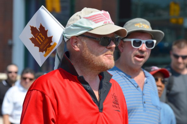Men walking in a labour day parade. They are wearing baseball caps. One of them has a small union flag tucked into the back of his cap. transit workers local 113