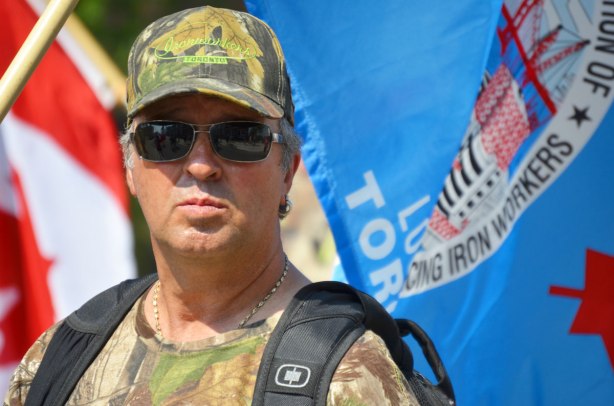 I man from the shoulders up. He is wearing a camo like t-shirt and hat with the ironworkers union label, and he is standing in front of a blue ironworkers flag. 