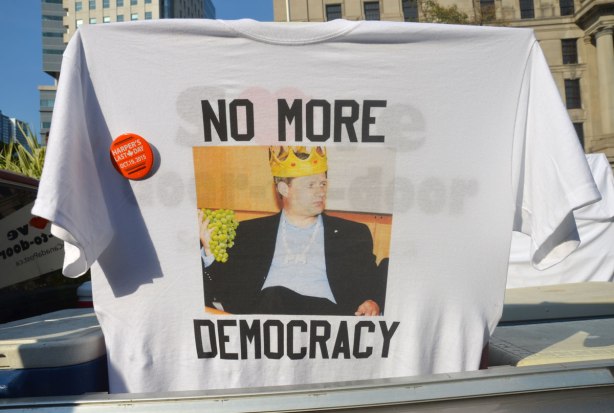 A t-shirt hanging in the window of a car. It has a picture of Stephen Harper wearing a crown and the words No More Democracy. There is a button on the t-shirt that says Harper's last day, 19th October 2015. 