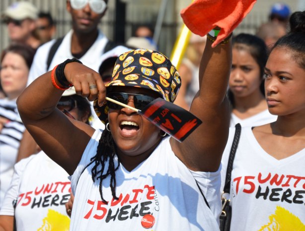 A black woman wearing a brightly coloured hat is waving a small sign that says I am not alone. She is walking in a labour day parade with a group from local 75
