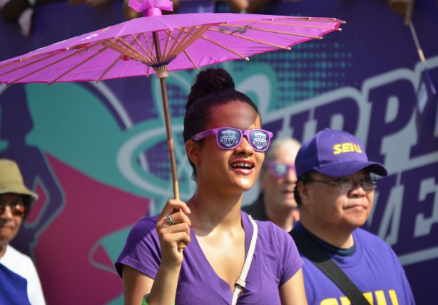 A woman carries a purple parasol as she walks in a labor day parade. She is wearing purple sunglasses that say Agent of Change on them 