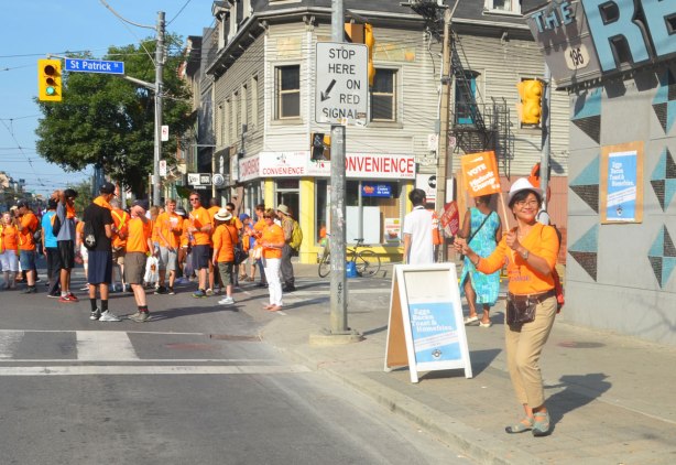 An Asian woman holds two small NDP flags as she stands on a sidewalk on Queen St. West by the Rex bar as she watches a labour day parade 