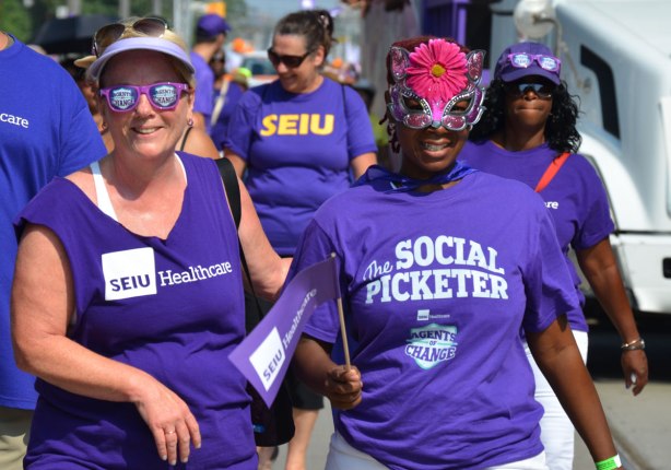 Four women in purple t-shirts from SEIU healthcare union walk in a labour day parade. One woman is wearing sunglasses with agents of change written on them. Another woman has the words The Social Picketer written on the front. 