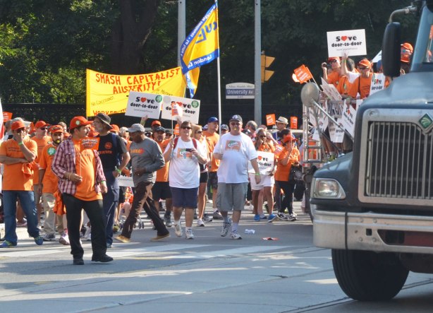 A group walks in a parade beside a large truck that is carrying more people on the back of it. They are carrying placards and banners in support of Canada Post. There is also a yellow banner that says Smash Capitalist Austerity 