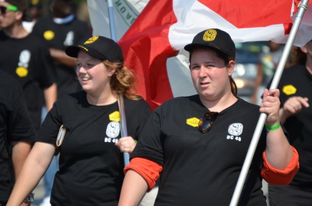 Two women in black caps and T-shirts are walking in a labour day parade. One of them is carrying a Canadian flag