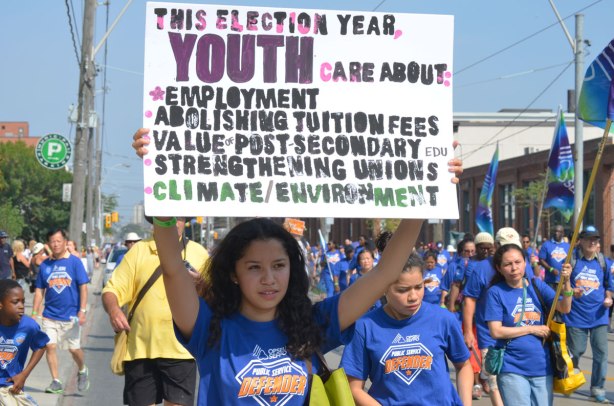 A young woman is holding up a sign about youth cares about and then it lists a few things like employment, tuition fees, unions, climate and the environment, at a Labour day parade