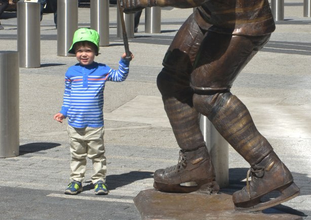 A little boy stands beside a statue of a hockey player where only the legs and skates of the player are in the picture. The boy is holding onto the end of the player's hockey stick