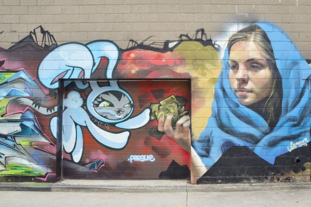 Street art on a wall in an alley, A Jarus woman with a blue shawl around her shoulders and a persue rabbit reaching for money that the woman is holding in her hand. 