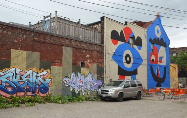 vacant lot, with a grey van parked in it, two pieces of street art on a wall as well as a large two storey mural 