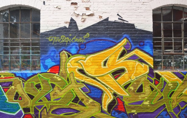 graffiti in yellows and greens on a blue background, on a wall in an alley, between two windows with metal grilles, signed BSM Crew