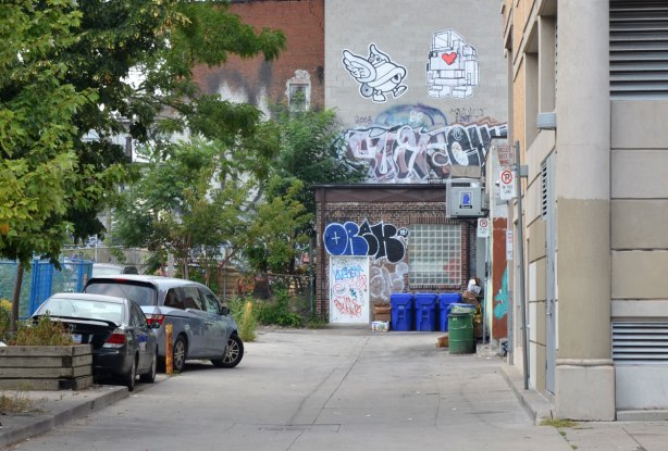 looking towards the end of an alley that has a large wheatpaste 3D lovebot high up on it. Beside lovebot is another wheatpaste 