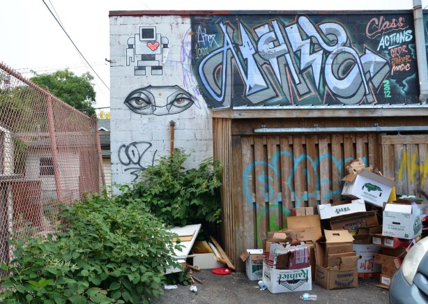 A lovebot and a pair of anser eyes on a wall in an alley. Lots of empty cardboard boxes on the ground by the wall.