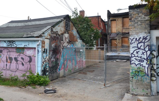 In an alley, the back of a house is being renovated, all the windows are boarded up and some of the exterior brick has been removed. There is a metal gate across the back of the property. The garage to the left is covered with graffiti.