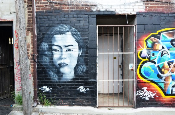 a wall with a painting of a woman's head on the left (in grey tones), a door with metal bar gate in the middle, and part of a tag like graffiti piece on the right.