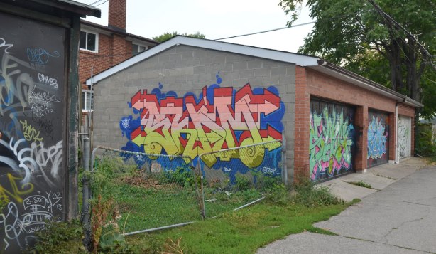 Newer garage in an alley with a crooked metal wire gate beside it. The garage has graffiti on the side wall and on both of its garage doors.