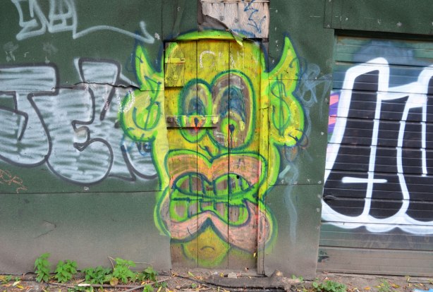 An old wood door on an alley building has been painted with a large yellowish green face with big pink lips and brown teeth