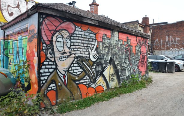 The front and side of a garage are covered with street art. The front is not very very visible in this picture but the side is. A man from the waist up with his finger pointing upwards is beside a picture of city buidings and a poser bunny.