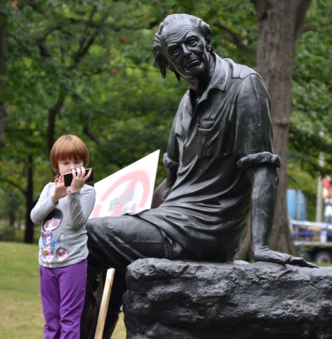 A statue of the Canadian poet Al Purdy sitting in a park. A girl looking at a smartphone is standing by its feet.