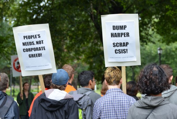Some people at a protest rally. Two young men are holding signs. One sign says Dump Harper Scrap CSIS. The other sign says People's needs not corporate greed.