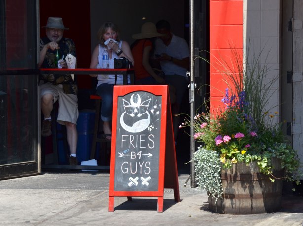 Chalkboard sign outside a restaurant that says Fries b 4 guys. Two people are inside the restaurant, sitting at a table and eating 
