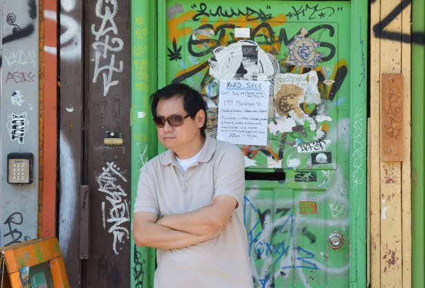 A man is standing in front of a green door that is covered with stickers, graffiti and bits of old posters 