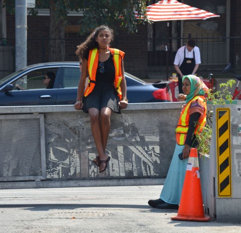 Two women in orange and yellow safety vests. One is sitting on a waist high barrier to keep cars off the street while the other woman is standing and leaning against it. 