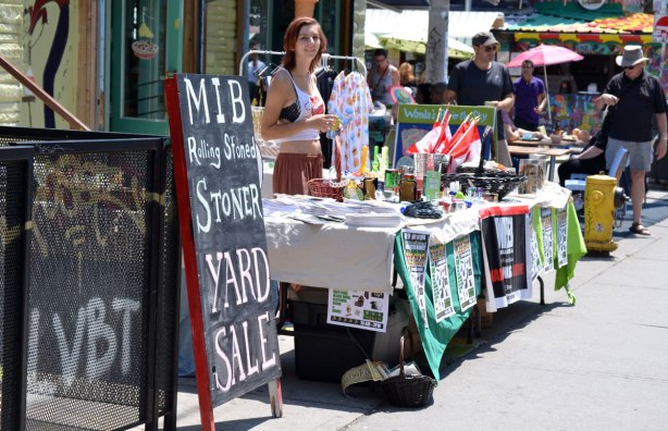 A woman is standing behind a table covered with things that she is selling, outdoors. A sign beside the table says MIB Rolling Stoned Stoner Yard Sale