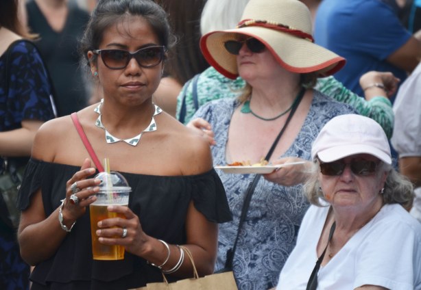 three women at an outdoor event. One is wearing sunglasses and a silver necklace. She is holding a cold drink in her hands. The other two women are older and are wearing hats. 