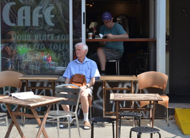 One man is sitting on a patio surrounded by a few empty tables and chairs. Right behind him is a man sitting at a table inside. The window is open so the man inside is visible 