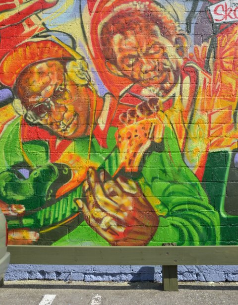 Part of a very colourful mural depicting various reggae musicians - A man wearing headphones and a baseball cap is playing a guitar. 