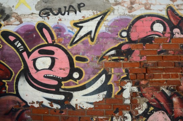 A street art painting is wearing away revealing the red brick underneath. A pink bunny is still partly visible. The word gwap is written above the rabbit's ears. 