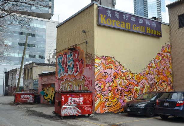 The back of the Korean Grill House restaurant, and the alley that its in. There is lots of street art on it. Two red garbage bins are also in the picture and they too have graffiti on them. 