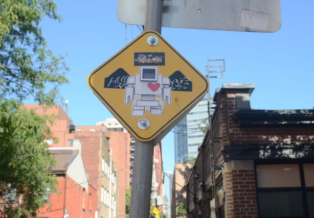 A yellow sign warning of speed bumps in the lane has a big lovebot sticker in the middle of it. Buildings in the background. 