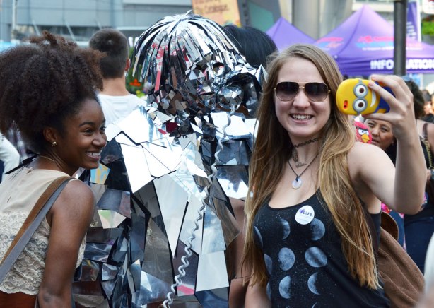 Two young women take a selfie with a phone that has a minion cover on it. They are at buskerfest in Toronto and they are posing with a character that is covered in pieces of shiny reflective material