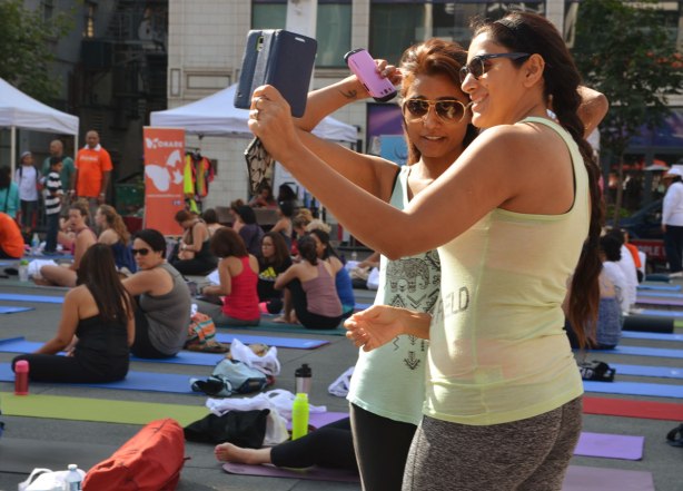 Two women taking a selfie at a yogathon, they are standing. Other people around them are sitting on mats, waiting for the event to begin. 
