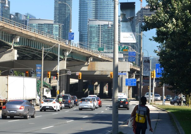 A woman is walking on a sidewalk, away from the camera, beside the Lakeshore Blvd in Toronto. There are a few cars on the road. There are skyscrapers in the background. The elevated highway, the Gardiner Expressway, is also in the picture. 