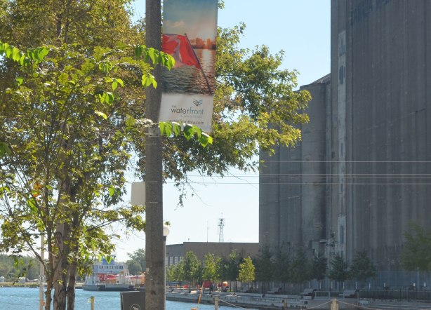 looking towards the waterfront. A banner with a picture of the Canadian flag is on a post by a tree in the foreground. The old silos for Canada Malting Company are in the background as is a boat moored beside the silos. 