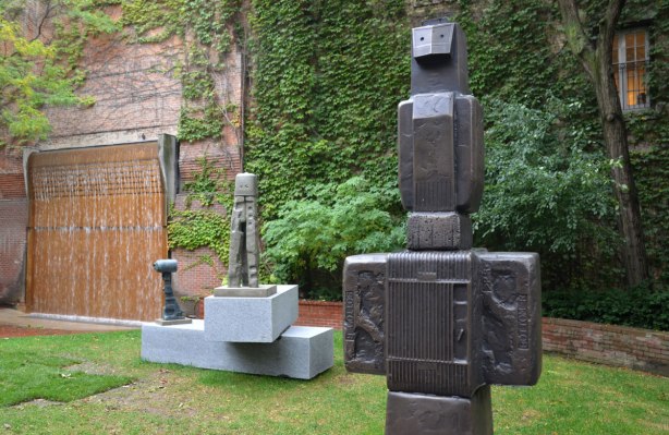 Three sculptures by An Te Liu in a sculpture garden, on pedestals in front of an ivy covered wall. 