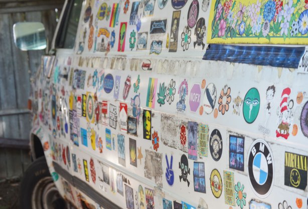 Drivers side of white chevy panel van, covered with stickers. 