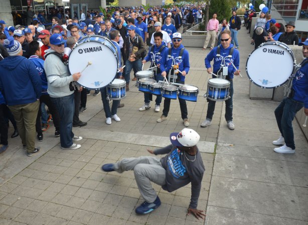 Before a Blue Jays baseball game at the ROgers Centre - a young man wearing a Jays grey T-shirt break dances in front of a5 man drum band who are all wearing blue Blue Jays T-shirts. There are many fans watching them play. 