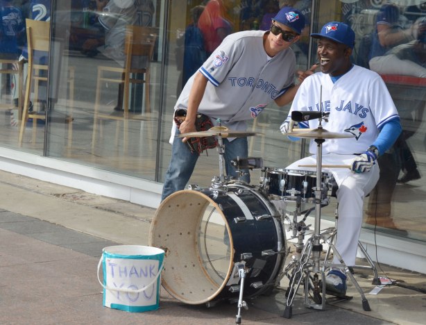 A man wearing a white Blue Jays uniform plays the drums while an onlooker poses beside him for a picture. 