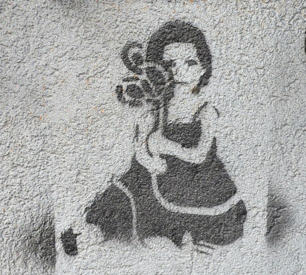 Stencil graffiti, black paint on grey wall, of a seated woman holding something in her hands. At first I thought she was holding a camera close to her face but I'm not sure. 
