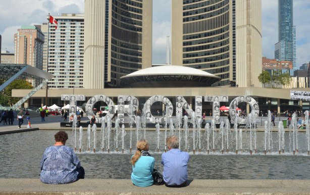 Three people sit on the bench beside the fountain at Nathan Phillips Square. Their backs are to the camera, they are looking towards the 3D Toronto sign that is covered with black and white photos for Inside Out project. Toronto city hall is in the background