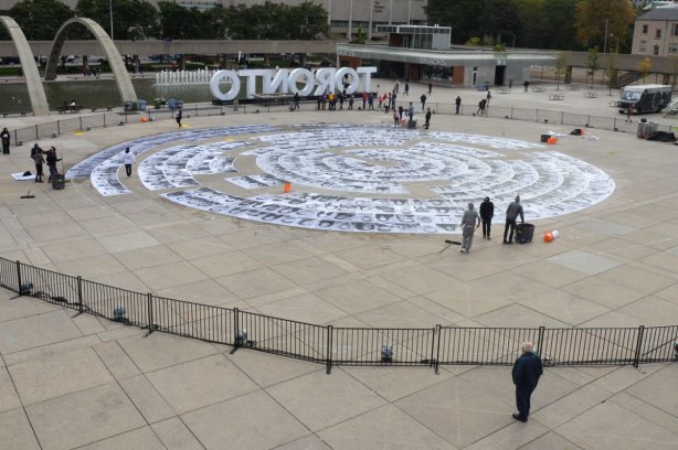 Concentric circles of black and white photos are being glued to the concrete at Nathan Phillips Square, overview, photo taken from the upper level. 