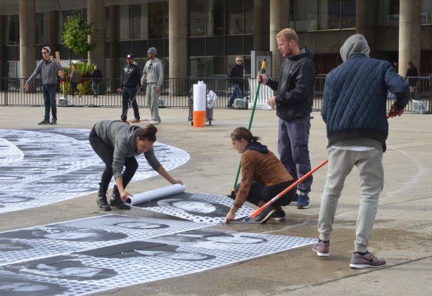 A group of young people are using brooms and sqeegees to glue blackand white photos of peoples faces onto the concrete of Nathan Phillips Square 