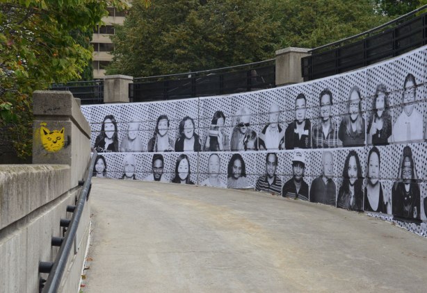 A wall alongside a sidewalk ramp is covered with black and white photos of people's faces. 