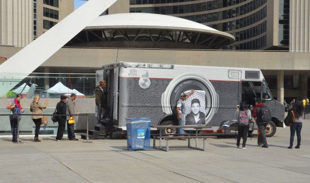 A picture of the mobile photoprinting booth that was used for the Inside Out global art project at Nathan Phillips Square. Some people are waiting in line to have their picture taken. 