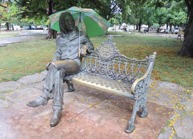 A statue of John Lennon sitting on a park bench in Havana. He's holding a green umbrella in the rain. 